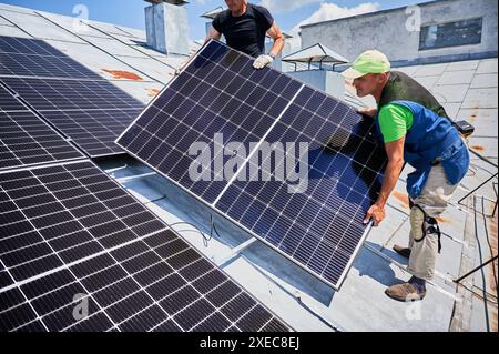 Ouvriers construisant le système de panneau solaire sur le toit métallique de la maison. Deux hommes installateurs portant le module solaire photovoltaïque à l'extérieur. Concept de production d'énergie alternative, verte et renouvelable. Banque D'Images