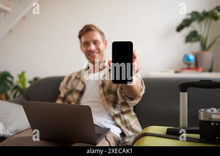 Portrait de beau jeune homme, montrant l'écran de smartphone, recommandant l'application mobile pour voyager ou réserver en ligne, assis esprit Banque D'Images