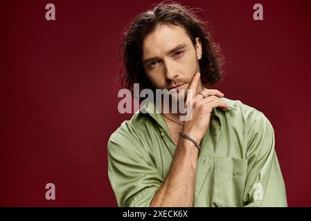 Un homme élégant aux lunettes et une chemise en soie verte éclatante prend la pose sur un fond rouge audacieux. Banque D'Images