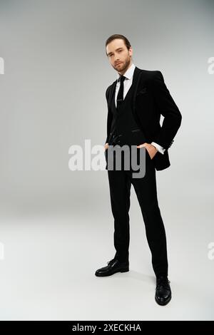 Un homme d'affaires barbu dans un élégant smoking pose avec confiance sur un fond de studio gris. Banque D'Images