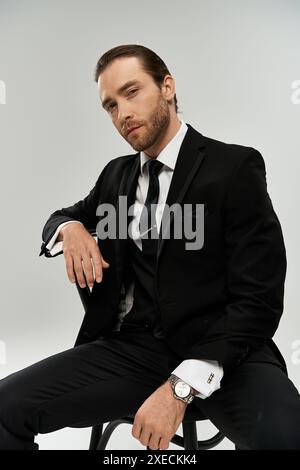 Un bel homme d'affaires barbu est assis sur une chaise dans un studio, regardant contemplatif dans son costume élégant sur un fond gris. Banque D'Images