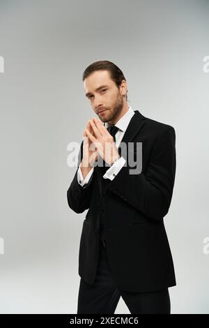 Un homme d'affaires barbu en costume et cravate se tient avec les mains attachées ensemble dans une pose réfléchie sur un fond gris. Banque D'Images