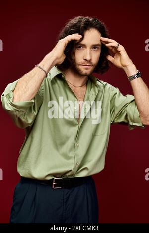 Un bel homme en lunettes et une chemise en soie verte prend une pose paisible sur un fond rouge vif. Banque D'Images