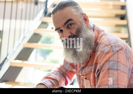 À la maison, homme senior avec une longue barbe assis dans les escaliers, regardant la caméra Banque D'Images