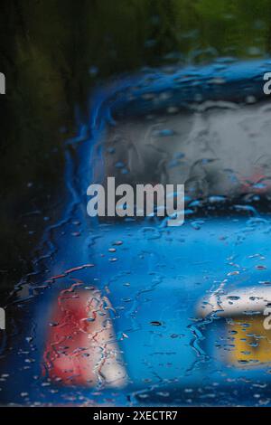 Une voiture bleue vide garée sur le bord de la route, vue à travers un pare-brise couvert de pluie. Banque D'Images