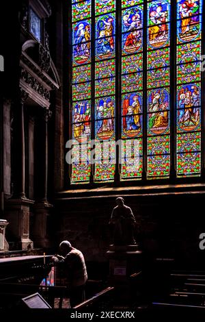 Un homme prie près des vitraux de la Collégiale Saint Aubin, Guérande, France. Les fenêtres colorées représentent des scènes religieuses. Banque D'Images