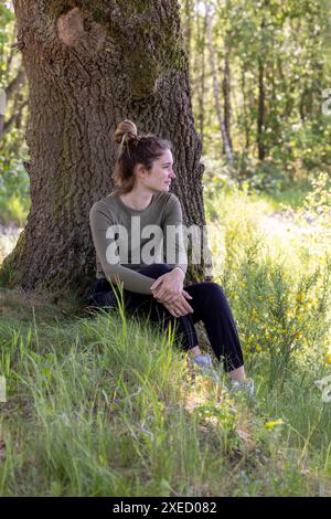 Femme réfléchie assise par un vieil arbre dans une forêt luxuriante Banque D'Images