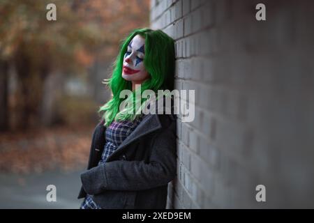 Portrait d'une fille aux cheveux verts en robe acclamée avec maquillage de joker sur un mur de brique flou d'arrière-plan. Banque D'Images