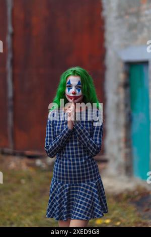 Portrait d'une fille aux cheveux verts en robe à l'entaille avec maquillage de joker sur un fond de mur en métal rouillé. Banque D'Images