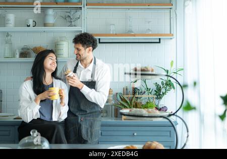 Un mari porte un tablier en cuisinant le dîner avec sa femme. Le duo a pris une pause pour siroter un café et déguster le pain complet. Banque D'Images