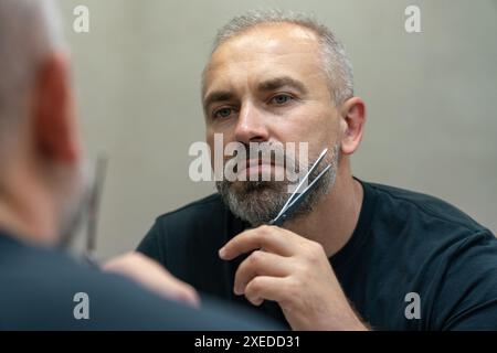 Portrait d'un beau homme d'âge moyen coupant sa barbe avec des ciseaux.Autosoins pendant l'isolement en quarantaine Banque D'Images