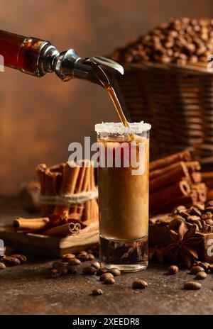 La liqueur de café est versée d'une bouteille dans un verre avec de la crème. Des grains de café, des bâtonnets de cannelle et de l'anis sont éparpillés sur la table. Banque D'Images