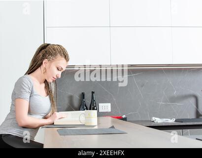 La jeune femme est assise à son comptoir de cuisine moderne, prenant une pause du travail pour lire un livre et profiter d'une tasse de café Banque D'Images