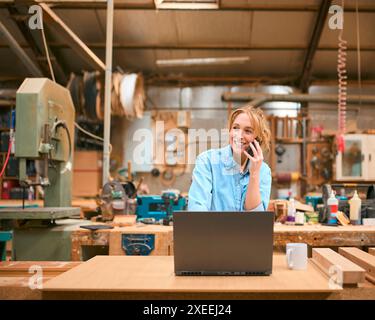 Menuisier féminin travaillant dans l'atelier de menuiserie parlant sur le téléphone portable et utilisant l'ordinateur portable Banque D'Images