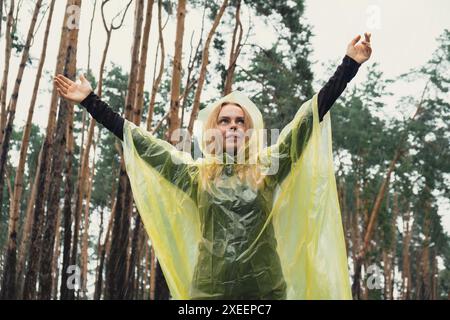 Femme souriante en imperméable jaune marchant dans la forêt d'automne profitant du temps pluvieux à l'extérieur. Touriste femme découvre le parc dans la pluie Banque D'Images