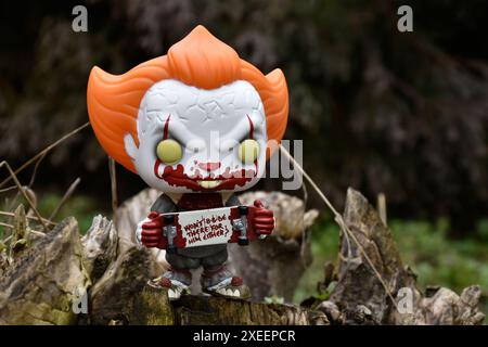 Funko Pop figurine d'action de Pennywise mal clown avec sanglant skateboard du film d'horreur il. Moody, sombre, effrayant, forêt vert foncé, souche d'arbre. Banque D'Images