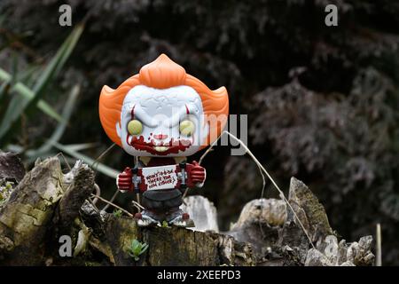 Funko Pop figurine d'action de Pennywise mal clown avec sanglant skateboard du film d'horreur il. Moody, sombre, effrayant, forêt vert foncé, souche d'arbre. Banque D'Images