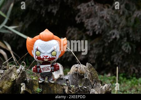 Funko Pop figurine d'action de Pennywise mal clown avec sanglant skateboard du film d'horreur il. Moody, sombre, effrayant, forêt vert foncé, souche d'arbre. Banque D'Images