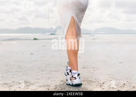 Gros plan des jambes d'une femme élancée dans une robe blanche du dos marchant dans le désert vers des montagnes lointaines Banque D'Images