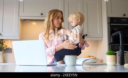 Stressée mère de travail avec jeune fils à la maison dans la cuisine en utilisant un ordinateur portable comme garçon joue avec son téléphone Banque D'Images