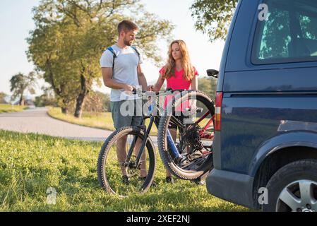 Le jeune homme décharge les vélos de montagne électriques, pour lui et sa petite amie, les soulevant de la galerie d'attelage du véhicule. Banque D'Images