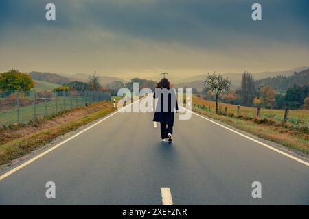 Une jeune femme au manteau noir marche seule le long d'une route asphaltée lisse, entourée d'arbres jaunes et d'herbe. Ciel dramatique et montagnes dans le backgro Banque D'Images