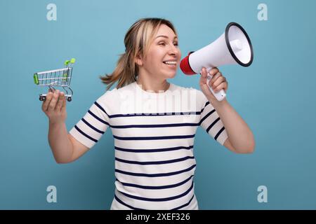 Énergique blonde jeune femme attrayante dans la tenue décontractée annonce la vente à l'aide de haut-parleur tenant le chariot d'épicerie sur fond bleu Banque D'Images