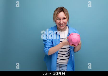 Blonde jeune femme attrayante en tenue décontractée avec un sourire tenant une tirelire avec des économies dans ses mains sur un fond bleu w Banque D'Images