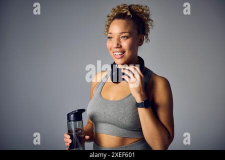 Studio shot de femme dans des vêtements de fitness de gymnastique avec casque sans fil et bouteille d'eau sur gris Banque D'Images