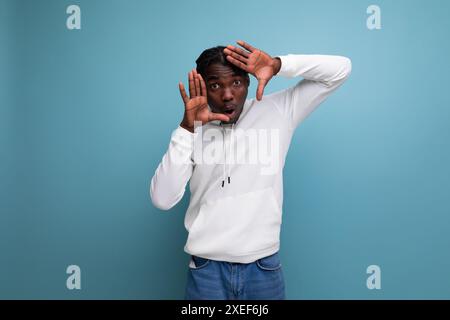 Jeune homme américain actif avec des dreadlocks dans un pull blanc inspiré montre sa main sur le côté sur un fond bleu Banque D'Images