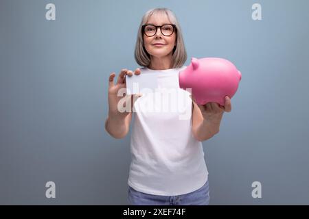 Compte épargne. femme mature avec les cheveux gris garde ses économies sur une carte de crédit et dans une tirelire sur un fond de studio avec Banque D'Images