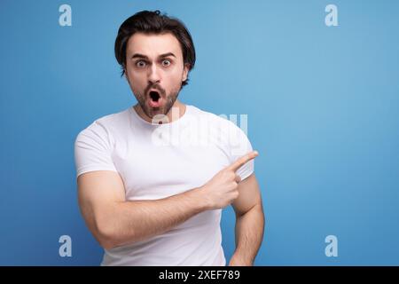 Choqué par surprise brune jeune homme dans un t-shirt blanc avec biceps sur un fond de studio avec copyspace Banque D'Images