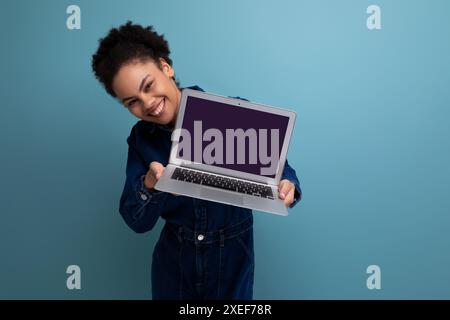 Jeune femme d'affaires hispanique joyeuse avec les cheveux moelleux habillé en costume en denim bleu montre l'écran d'ordinateur portable avec maquette Banque D'Images