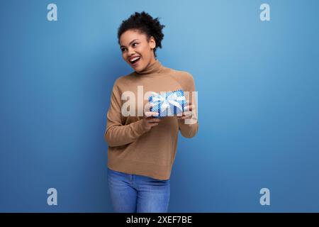 Jeune femme joyeuse joyeuse avec la peau foncée et les cheveux moelleux avec boîte-cadeau sur fond isolé avec espace de copie Banque D'Images
