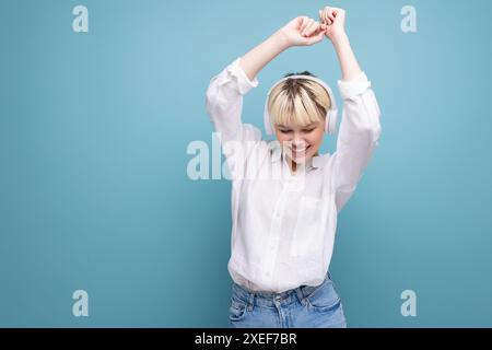 Jeune jolie étudiante blonde vêtue d'une blouse blanche écoute sa musique préférée au casque sans fil sur le bac Banque D'Images