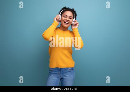 Jeune femme d'affaires hispanique joyeuse aux cheveux bouclés noirs écoutant de la musique dans des écouteurs blancs Banque D'Images