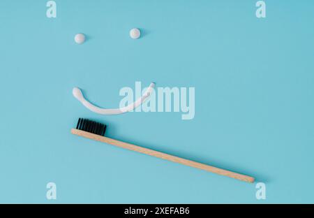 Brosse à dents en bambou organique et dentifrice médical blanc faisant visage de sourire sur fond bleu pastel. Concept dentaire minimal. Dents saines et blanches. Banque D'Images