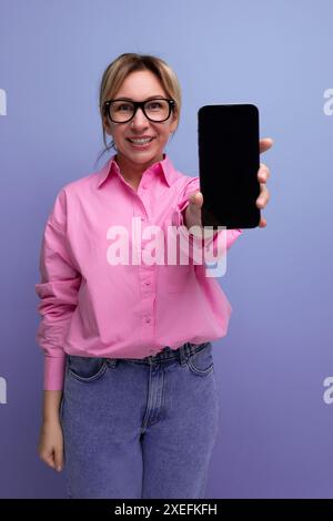 Jeune femme dirigeante blonde blonde réussie dans une blouse rose et des lunettes montre un écran de smartphone avec une maquette pour insertin Banque D'Images