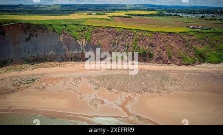 Vue aérienne d'une falaise côtière avec une végétation verdoyante au sommet, surplombant une plage de sable avec des rochers éparpillés et la mer. Les terres agricoles et les bâtiments sont vi Banque D'Images