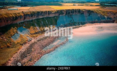 Vue aérienne d'une falaise côtière avec une plage de sable et une eau bleu clair. Le paysage comprend des champs verts au sommet de la falaise et un rivage rocheux. Banque D'Images
