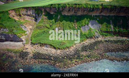 Vue aérienne d'une falaise côtière avec végétation verte, rivage rocheux et eau bleue claire. La falaise montre des signes d'érosion et de glissements de terrain. Banque D'Images