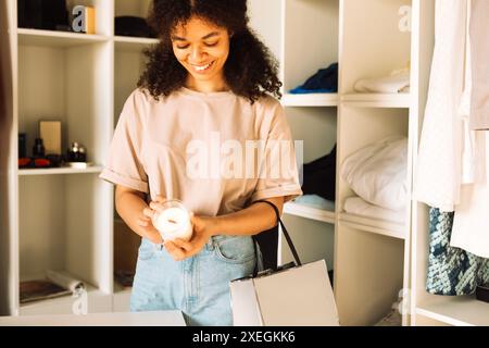 Charmante adolescente de race mixte choisir la bougie en magasin. Attrayante jeune femme africaine dans des vêtements décontractés tient une bougie parfumée Banque D'Images