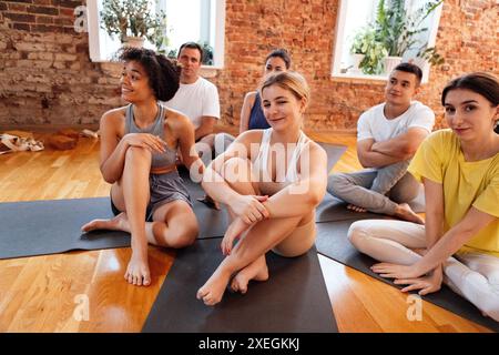 Les gens souriants multiraciaux s'assoient sur des tapis après avoir fait du yoga au club de fitness. Groupe de jeunes athlètes de différents âges et nationalit Banque D'Images