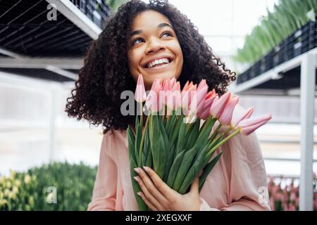 Belle fille d'ethnicité africaine souriante romantique tenant un bouquet de tulipes roses dans la serre. Gros plan de jeune afro féminin Banque D'Images