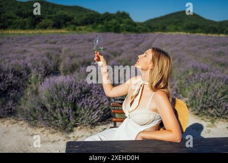 Jeune femme blonde en robe blanche est assise sur une chaise parmi le champ de lavande et tient un verre de vin mousseux Banque D'Images