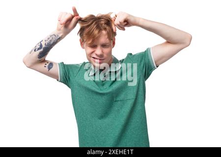 Jeune homme européen élégant avec les cheveux roux avec un tatouage sur son avant-bras porte un t-shirt vert touche ses cheveux Banque D'Images