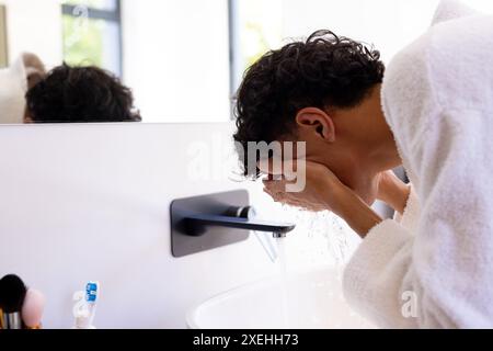 Se laver le visage dans la salle de bain, jeune homme portant une robe blanche devant le miroir Banque D'Images