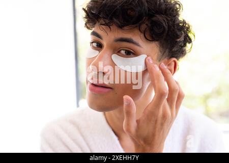 Application de patchs sous les yeux, jeune homme en peignoir appréciant la routine de soins de la peau à la maison Banque D'Images