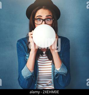 Choqué, femme et ballon avec soufflage en studio pour la célébration, le plaisir et le plaisir sur fond bleu. Organisateur d'événements, lunettes et personne avec Banque D'Images