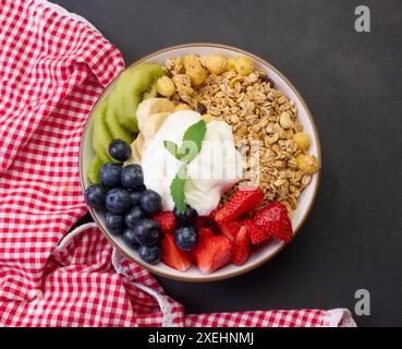 Petit déjeuner sain. Granola avec fraises, kiwi, banane et bleuets dans une assiette ronde sur une table noire. Banque D'Images